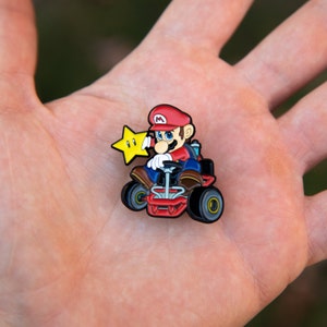 Pu&ograve; includere: Uno spillo con un personaggio di Mario Kart rosso e blu con una stella gialla. Il personaggio guida un kart rosso con ruote nere.