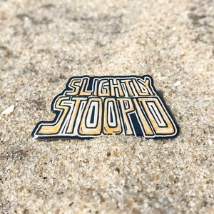 Könnte beinhalten: Rechteckiger Emaille-Pin mit dem Schriftzug "SLIGHTLY STOOPID" in einer stilisierten Schriftart. Der Text ist schwarz umrandet und mit einer gelb-goldenen Farbe gefüllt. Der Pin befindet sich auf einem sandigen Hintergrund.