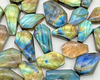 May include: A collection of polished labradorite gemstones in various shapes. The stones display a mix of colors, including blues, greens, and golds, with iridescent flashes. The gemstones are arranged on a white surface.