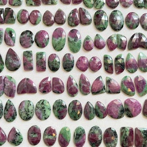 May include: A collection of faceted ruby zoisite gemstones in various shapes including ovals, teardrops, and rectangles. Each stone displays a unique combination of deep magenta-pink ruby inclusions contrasted against vibrant green zoisite mineral matrix.