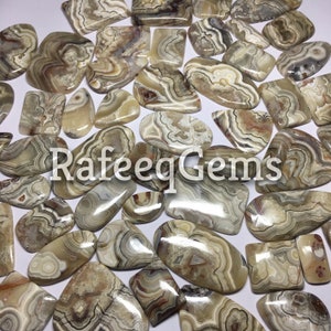 May include: A collection of polished agate gemstones in various shapes and sizes, showcasing natural patterns of beige, brown, and gray. The gemstones are scattered across a white surface, with the text "RafeeqGems" overlaid on the image.