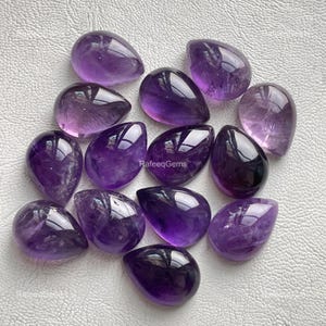 Puede incluir: Una colección de trece piedras preciosas de amatista en forma de lágrima en varios tonos de púrpura, que van desde el lavanda claro hasta el violeta intenso. Cada piedra pulida tiene una superficie lisa y reflectante con el texto RafeeqGems.