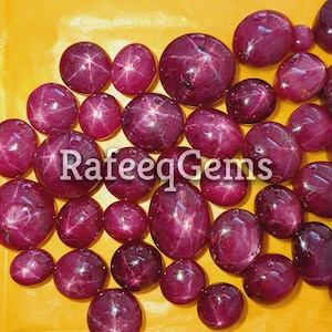 May include: A collection of polished, deep magenta star rubies in various sizes, each displaying a distinct six-rayed star effect on its smooth, cabochon-cut surface. The text RafeeqGems is overlaid in white across the center of the gemstones.