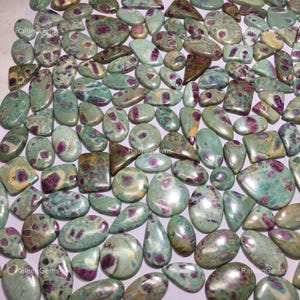 Op de afbeelding: Een verzameling groene en roze ruby zoisiet cabochons. De stenen zijn in verschillende vormen en maten, met een mix van ovale, druppelvormige en rechthoekige vormen. De stenen hebben een groene basis met roze en witte insluitsels.