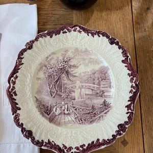 May include: A white ceramic dinner plate with a purple floral design. The plate has a raised rim with a scalloped edge. The center of the plate features a purple and white landscape scene with a bridge and a waterfall.