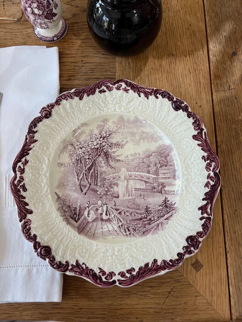 May include: A white ceramic dinner plate with a purple floral design. The plate has a raised rim with a scalloped edge. The center of the plate features a purple and white landscape scene with a bridge and a waterfall.