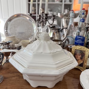May include: A white ceramic tureen with an octagonal base and a matching lid featuring a decorative finial. The tureen has a wide rim and a handle extending from the side, showcasing a clean, solid white finish throughout its structured design.