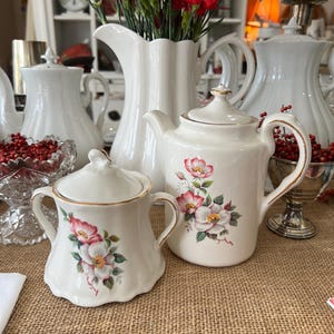 Op de afbeelding: Witte keramische theeset met bloemenmotieven. De set bevat een theepot, een roomkan en een suikerpot. De theepot heeft een gouden rand en een bloemenmotief met roze rozen. De roomkan en de suikerpot hebben een vergelijkbaar bloemenmotief.