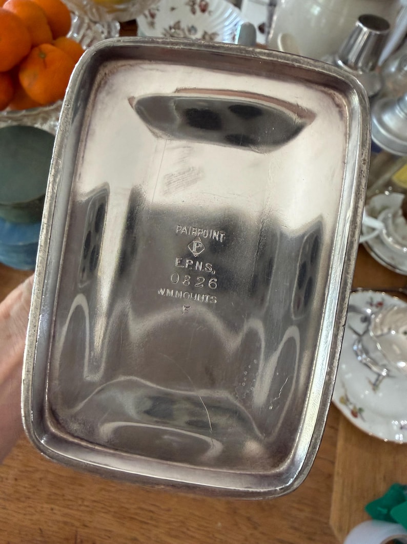 May include: A rectangular silver-colored tray with rounded corners. The tray has a slightly raised rim and a handle-like indentation on one side. The bottom of the tray is stamped with the words "PAIRPOINT", "E.P.N.S.", "0826", and "WM.MOUNTS".