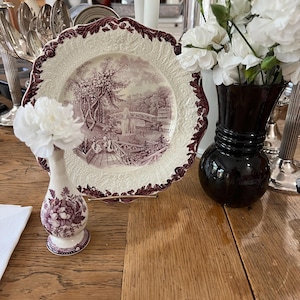 May include: A vintage porcelain plate with a burgundy and white floral design, featuring a scene of a bridge and a fountain. The plate is on a wooden table with a small white vase with burgundy floral accents and a black vase with white flowers.