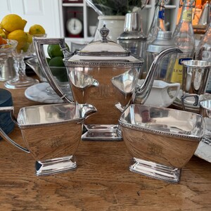 May include: A silver tea set with a teapot, creamer, and sugar bowl, all with ornate detailing and a monogram. The set is displayed on a wooden table, with other items in the background.