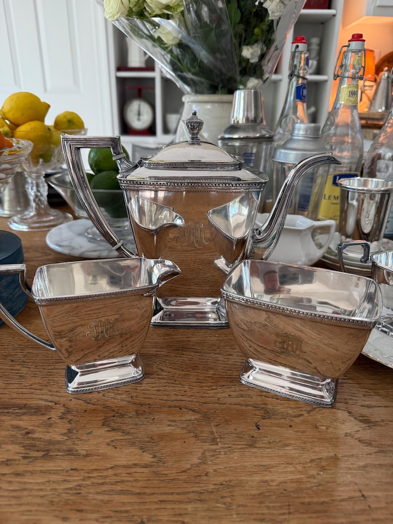 May include: A silver tea set with a teapot, creamer, and sugar bowl, all with ornate detailing and a monogram. The set is displayed on a wooden table, with other items in the background.