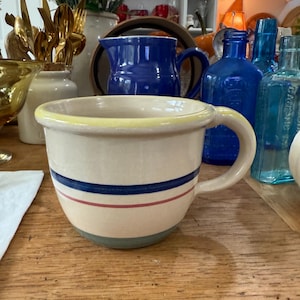 May include: Cream-colored ceramic mug with a yellow rim and a handle. The mug features two horizontal stripes: a dark blue stripe above a red stripe. The base of the mug is light blue. Other vintage items are in the background.