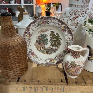 May include: A vintage brown and white ceramic jug with a floral design, a brown wicker bottle, a white ceramic bottle, and a brown and white ceramic plate with a floral design and a countryside scene. The items are arranged on a wooden crate with the text "V-A-N" and "BRIGHTON" printed on it.
