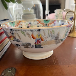 May include: A white porcelain bowl with a gold rim and a colorful Asian-inspired design. The bowl features figures in traditional clothing and a landscape scene.