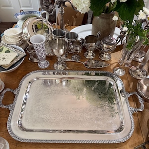 May include: A large rectangular silver serving tray with ornate engraved scrollwork patterns on the surface and a twisted rope-style border. The tray features two decorative handles on the sides and has a polished, reflective metallic finish.