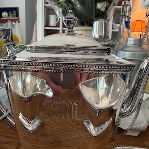 May include: A silver tea set with a wooden base. The set includes a teapot with a decorative lid, and a tray. The teapot has a monogram. The set is likely vintage and suitable for serving tea.