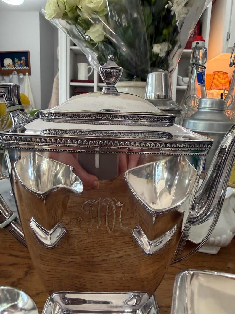 May include: A silver tea set with a wooden base. The set includes a teapot with a decorative lid, and a tray. The teapot has a monogram. The set is likely vintage and suitable for serving tea.