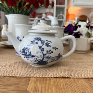 May include: A small white teapot with a blue and white floral design. The teapot has a lid and a spout. It is sitting on a brown burlap surface.