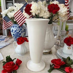 Könnte beinhalten: Eine weiße Keramikvase mit einer hohen, schlanken Form und einem ausgestellten Fuß. Die Vase ist mit einem Strauß weißer und roter Nelken sowie blauen Distelblüten gefüllt. Die Vase steht auf einem Tisch mit einer Tischdecke aus Sackleinen.