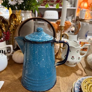 May include: A vintage blue enamelware coffee pot with white speckles. The pot has a lid, a handle, and a spout. The pot is sitting on a wooden table. The pot is part of a collection of vintage kitchenware.