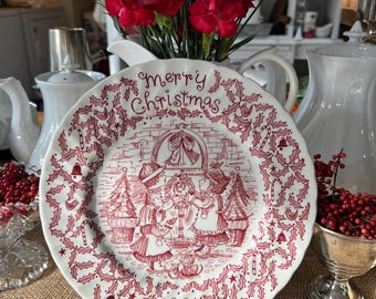 May include: A white ceramic Christmas plate with a red and white design. The plate features a scene of children singing carols in front of a fireplace. The text "Merry Christmas 2001" is printed on the plate.