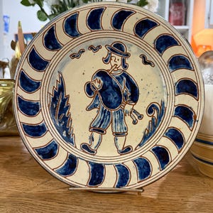 Puede incluir: Un plato redondo de cerámica pintado a mano con un diseño azul y blanco. El plato presenta una ilustración central de una persona con ropa azul tocando un instrumento musical, rodeada por un borde azul y blanco. El plato se exhibe sobre un soporte metálico.