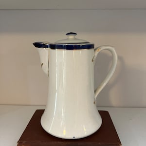 May include: White ceramic coffee pot with a blue and gold trim. The pot has a rounded body and a spout on one side. The lid has a blue trim and a gold band. The pot is sitting on a brown book.