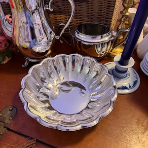 May include: A silver-plated tea service set consisting of a tall pitcher with a curved handle, a small open sugar bowl, and a shallow scalloped bowl. The pieces feature a polished, reflective metallic finish and ornate, fluted edge detailing.