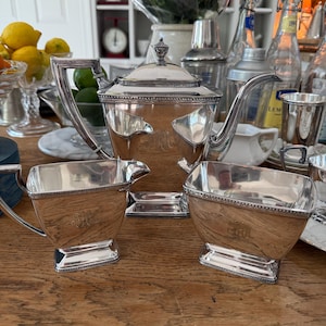 May include: A silver tea set with a teapot, creamer, and sugar bowl, all with ornate detailing and a monogram. The set is displayed on a wooden table, with other items in the background.