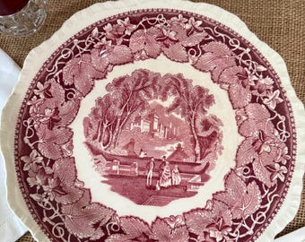 May include: A white and burgundy plate with a scalloped edge. The plate features a burgundy illustration of a landscape scene surrounded by a border of leaves and vines. Other serving dishes are in the background.