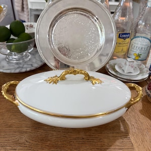 Puede incluir: Fuente ovalada blanca con detalles dorados sobre una mesa de madera. La fuente tiene un asa dorada y un borde dorado. Un plato plateado y otros platos están en el fondo.