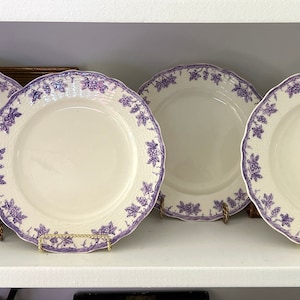 May include: Four white ceramic dinner plates with a purple floral design. The plates are on a white shelf.