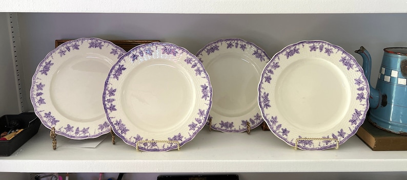 May include: Four white ceramic dinner plates with a purple floral design. The plates are on a white shelf.