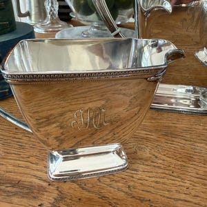 May include: A silver-plated serving set, including a creamer, sugar bowl, and tray, is displayed on a wooden surface. The creamer features a spout and handle, with an engraved monogram. The set has a decorative beaded border.