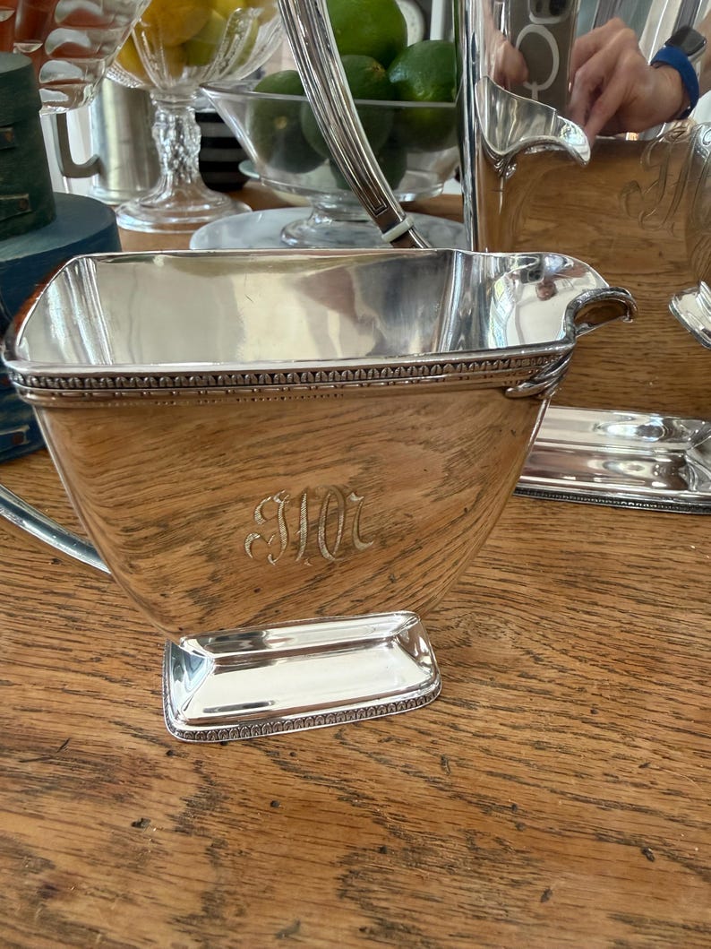 May include: A silver-plated serving set, including a creamer, sugar bowl, and tray, is displayed on a wooden surface. The creamer features a spout and handle, with an engraved monogram. The set has a decorative beaded border.
