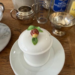 May include: A white ceramic dish with a lid, topped with a decorative fruit and leaf design, sits on a stack of white plates. Other items include a silver sugar bowl, a measuring glass, and a small silver bowl.