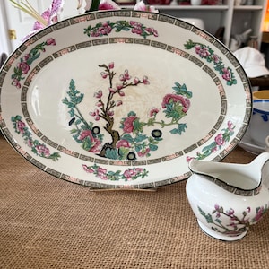 May include: An oval serving platter and a small creamer with a floral design. The platter has a border of pink flowers and a central design of a flowering tree. The creamer has a matching floral pattern.