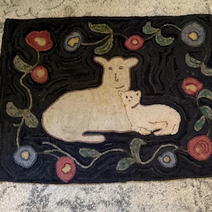 Op de afbeelding: Handgehaakte vloerkleed met een crèmekleurig lam en zijn jong op een donkere achtergrond. Het vloerkleed is versierd met kleurrijke bloemen en groene bladeren. Het vloerkleed is rechthoekig.
