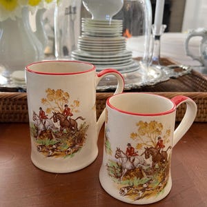 Puede incluir: Dos tazas de cerámica blanca con ribete rojo y un diseño de escena de caza. Las tazas muestran jinetes a caballo con perros corriendo a su lado.