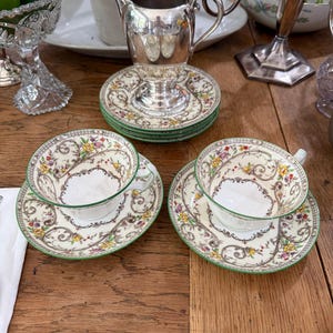 May include: A silver creamer jug and two porcelain teacups with matching saucers. The teacups and saucers feature a cream-coloured base with intricate brown scrollwork, green rims, and small yellow, red, and purple floral accents.