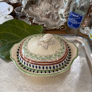 May include: A round ceramic butter dish with a lid featuring a cream-colored base, hand-painted green leaf garlands, brown bands, and blue vertical dashes. The lid is topped with a sculpted ceramic leaf handle and has scalloped edges.