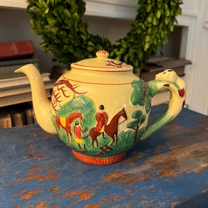Pode incluir: Uma chaleira amarela com uma cena de caça verde e marrom pintada no lado. A chaleira tem uma alça marrom e uma tampa marrom com uma pequena maçaneta.