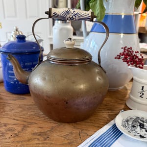 May include: Antique copper kettle with a brass spout and handle. The handle is decorated with a white and blue floral design. The kettle sits on a wooden surface, with other vintage items in the background.