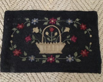 Puede incluir: Una alfombra negra de ganchillo con una cesta de color crema llena de flores. La cesta está rodeada de una corona de flores en tonos rojos, azules y crema.