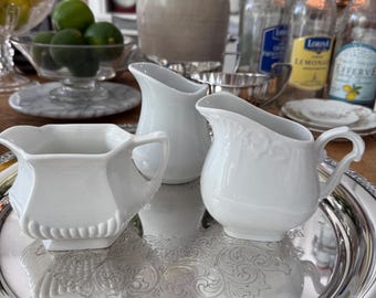 Może przedstawiać: Trzy białe ceramiczne dzbanki na śmietankę na srebrnej tacy z ozdobnymi detalami. Dzbanki mają różne kształty, jeden z kwadratową podstawą, a pozostałe z zaokrąglonymi korpusami. Taca jest okrągła z dekoracyjną krawędzią.
