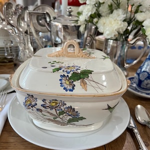 May include: A white porcelain serving dish with a floral design. The dish has a lid with a decorative handle. The dish is on a white plate and a wooden table.