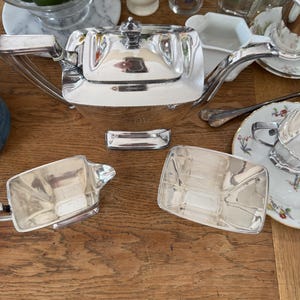 May include: A silver tea set, including a teapot and two rectangular serving dishes, sits on a wooden surface. The teapot has a square shape with a lid and a curved handle. Limes in a glass bowl are visible.