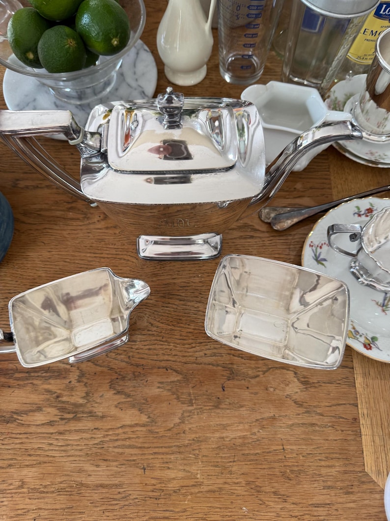 May include: A silver tea set, including a teapot and two rectangular serving dishes, sits on a wooden surface. The teapot has a square shape with a lid and a curved handle. Limes in a glass bowl are visible.