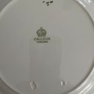 May include: White ceramic plate with a green and black mark that reads "CAULDON ENGLAND" and a crown above the text. The plate also has a small black mark with the letters "WX" and a number "0" below.